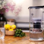Eine gläserne Wasserfilterkanne mit Filterkartusche steht auf einem Holztisch neben einem gefüllten Wasserglas und frischen Zitronen.