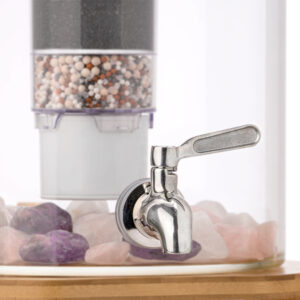 Nahaufnahme eines Edelstahl-Wasserhahns an einem Glas-Wasserspender, im Hintergrund eine mehrstufige Filterkartusche und am Boden Edelsteine.