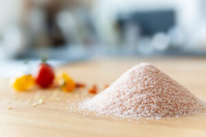Ein Haufen feines rosa Kristallsalz liegt auf einem Holzbrett, mit bunten Tomaten im Hintergrund.