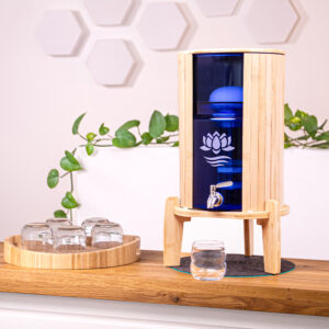Ein Wasserfilter mit Bambusgehäuse und blauem Glasbehälter steht auf einer Holztheke, daneben ein Tablett mit Gläsern.