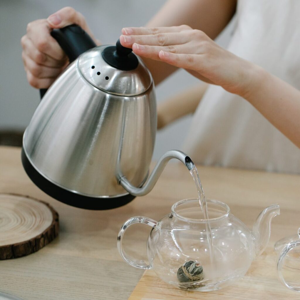 Eine Person gießt heißes Wasser aus einem Edelstahl-Wasserkocher mit Schwanenhals in eine gläserne Teekanne, um einen Teeball aufzubrühen.