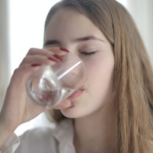 Eine junge Frau trinkt mit geschlossenen Augen genussvoll ein Glas klares Wasser.