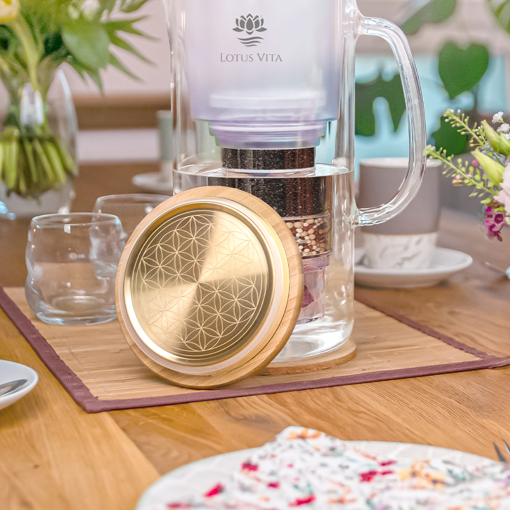 Eine stilvolle Glas-Wasserfilterkanne von Lotus Vita steht auf einem gedeckten Holztisch, davor liegt der goldene Bambusdeckel mit einer Gravur der Lebensblume.
