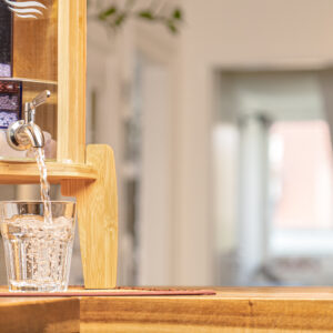 Ein Wasserfilter-Standgerät aus Bambus mit einem Zapfhahn aus Edelstahl befüllt ein Trinkglas mit frischem, klarem Wasser.