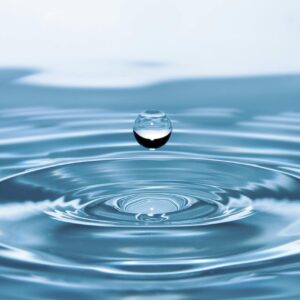 Ein klarer Wassertropfen schwebt über einer Wasseroberfläche und zieht sanfte, kreisförmige Wellen.