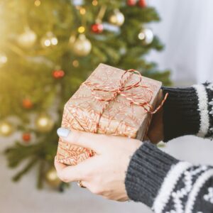 Eine Person in einem winterlichen Pullover hält ein liebevoll verpacktes Geschenk, im Hintergrund leuchtet ein geschmückter Weihnachtsbaum.