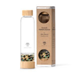 Eine Glas-Trinkflasche mit Bambusdeckel und einem separaten Bodenfach, gefüllt mit einer Mischung aus goldenen, schwarzen und türkisfarbenen Edelsteinen, steht neben ihrer Kartonverpackung.