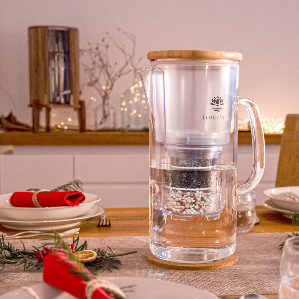 Eine Glas-Wasserfilterkanne mit Bambusdeckel steht auf einem festlich für Weihnachten gedeckten Holztisch, umgeben von Dekoration.