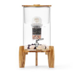 Ein stilvoller Glas-Wasserspender mit Deckel und Standfuß aus Akazienholz, ausgestattet mit einer sichtbaren Filterkartusche und dekorativen Edelsteinen am Boden.