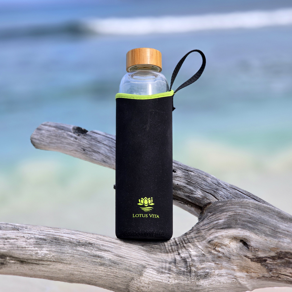 Eine Glas-Trinkflasche mit Bambusdeckel und schwarz-grüner Schutzhülle, die auf einem Stück Treibholz vor einer unscharfen Meereskulisse liegt.