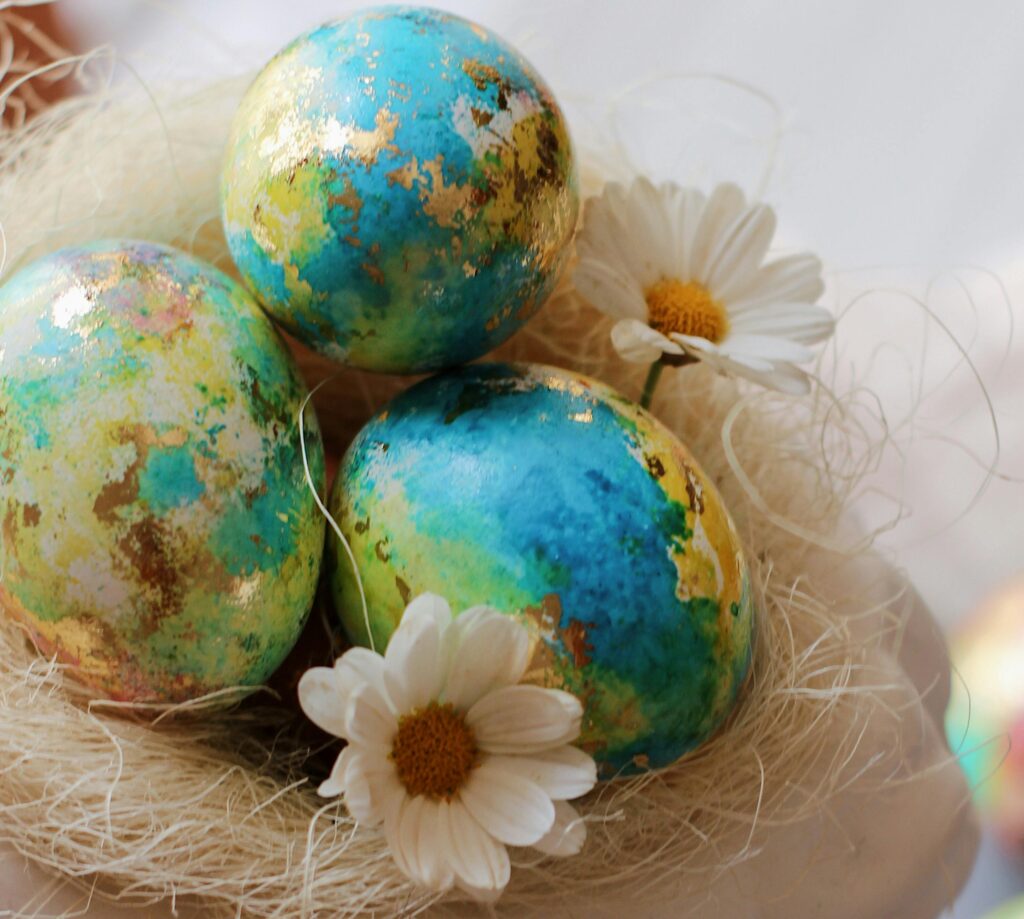 Kunstvoll in Blau-, Grün- und Goldtönen bemalte Ostereier liegen zusammen mit kleinen Gänseblümchen in einem stilvollen, weißen Osternest.