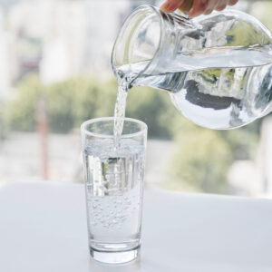 Eine Hand gießt klares, gefiltertes Wasser aus einer Glaskaraffe in ein Trinkglas.