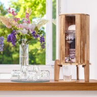 Ein Wasserfilter mit Akazienholz-Gehäuse steht auf einer Holztheke neben mehreren Gläsern, im Hintergrund ein sonniges Fenster mit Blumen.