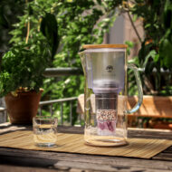 Eine Glas-Filterkaraffe mit Bambusdeckel und einem Glas gefilterten Wassers steht auf einem Holztisch auf einem sonnigen Balkon.